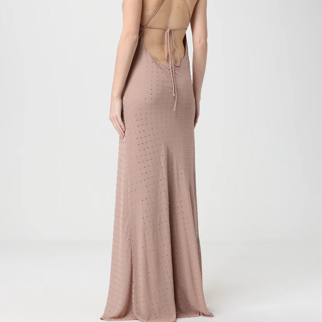 A woman shown from the back wearing a floor-length dusty blush pink dress with a strappy open back and tie detail, photographed against a clean light gray studio background.